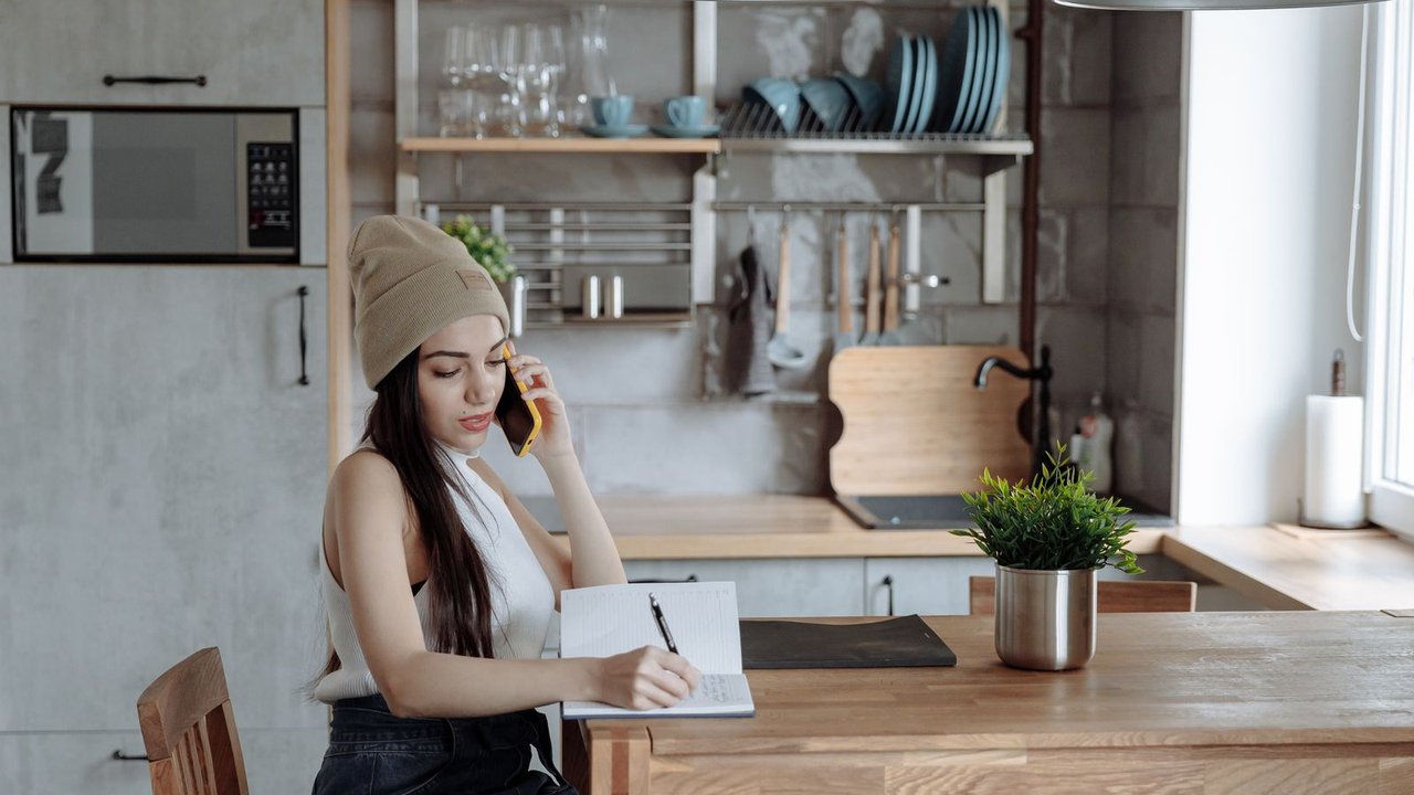 A woman at her kitchen table talking on the phone while making notes.