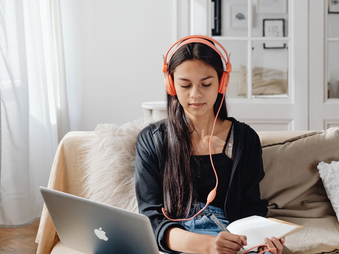 A woman on a sofa with headphones and a laptop, taking notes as she learns.