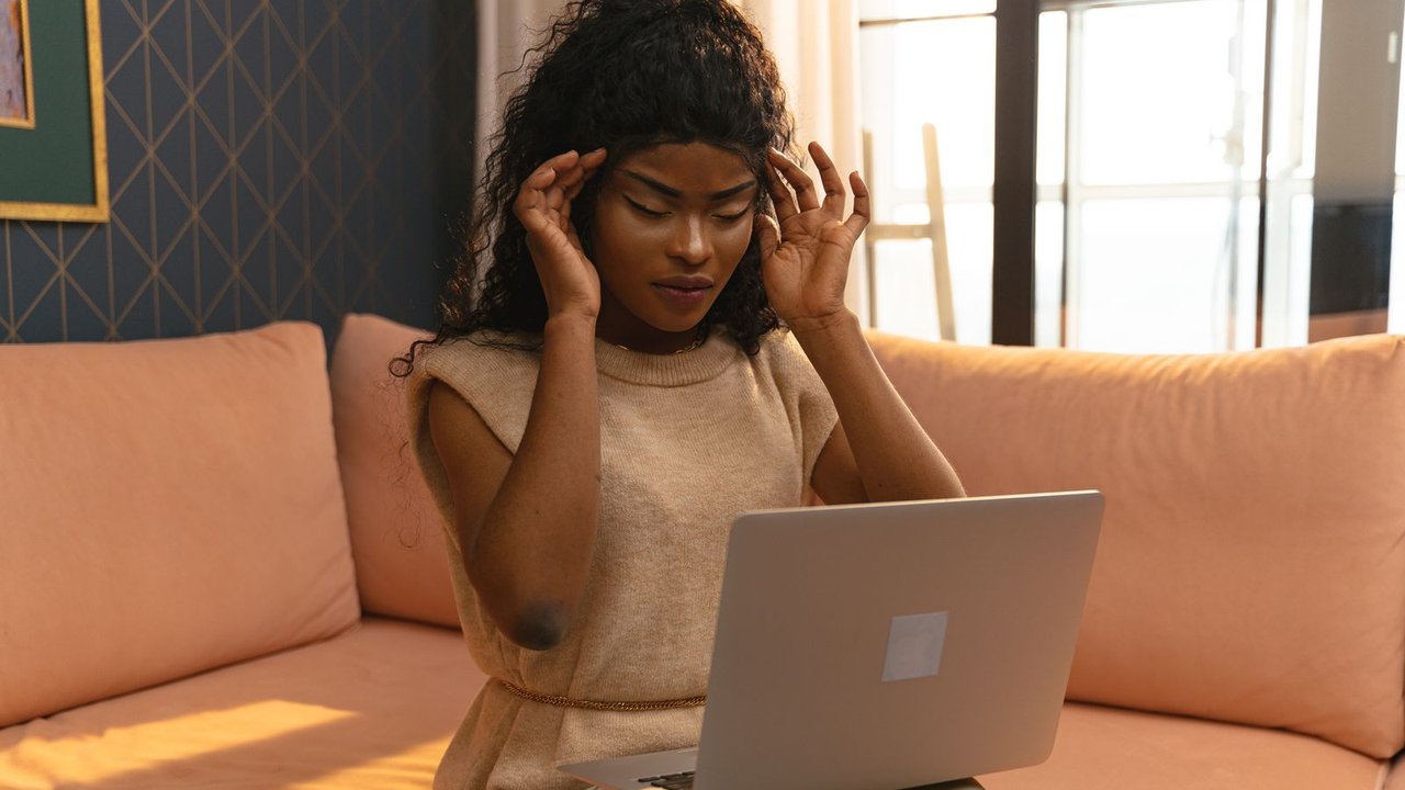A woman on a peach sofa with a laptop on her knees, pressing her fingers to her temples.